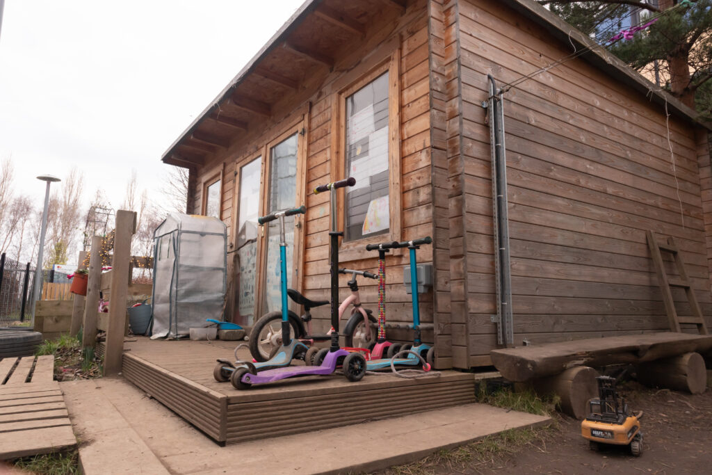 The hut in hidden garden area of the nursery. Scooters are parked outside the hut and there is a construction vehicle toy on the ground.