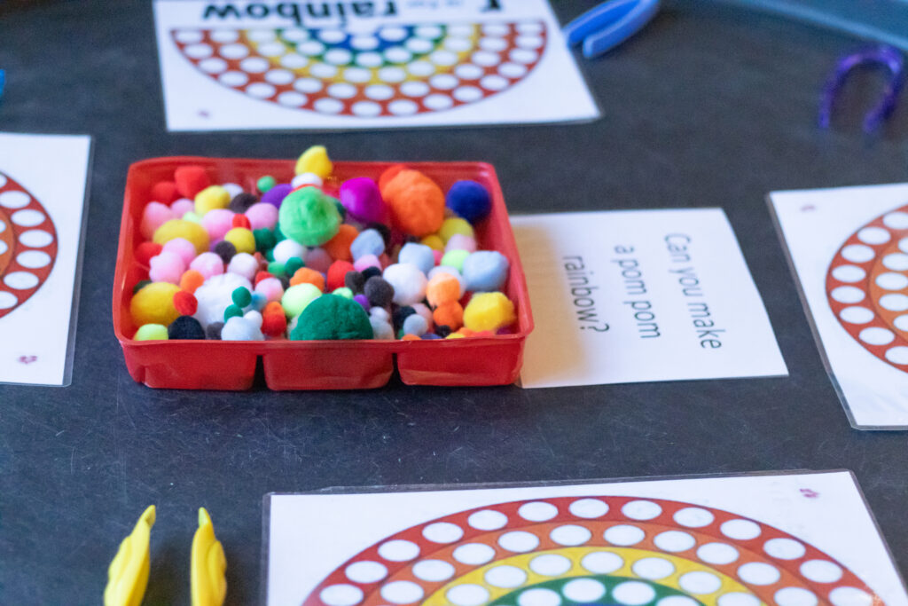 Pom poms of various sizes and colours in a tray. There is a sign next to the tray that says 'Can you make a pom pom rainbow?'.