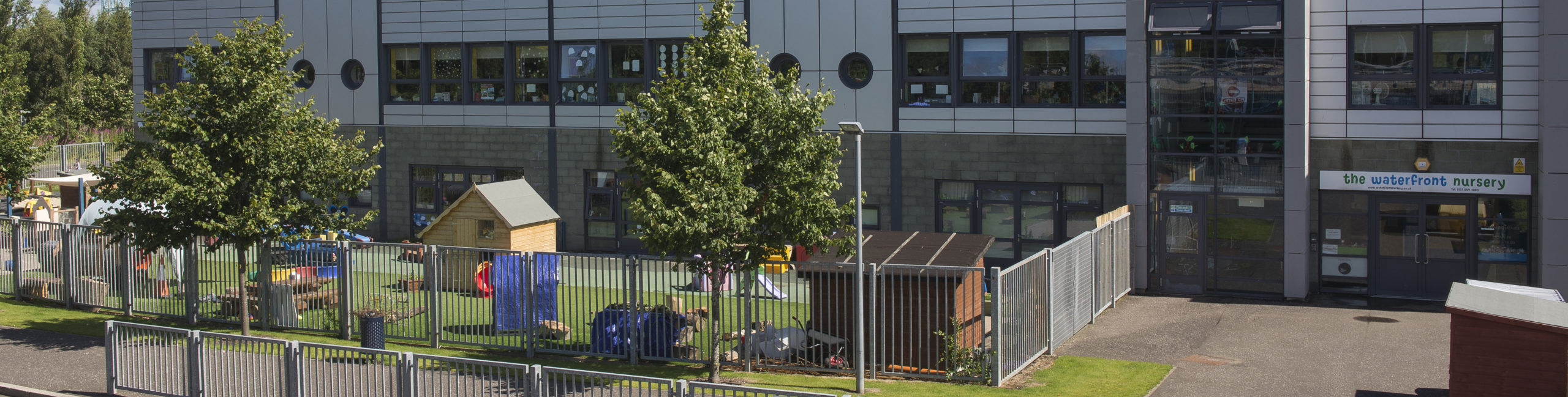 Outdoor view of the nursery building and the outdoor play area. There is a sign above the nursery entrance saying 'the waterfront nursery'.